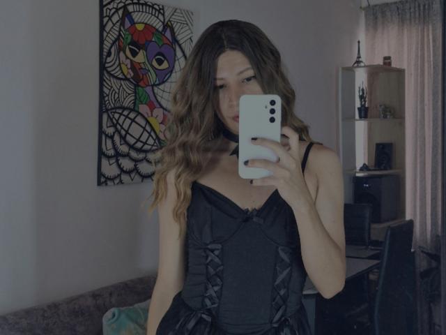 SanzJulie with wavy brown hair, wearing a black satin corset, holding a white phone, taking a mirror selfie, showing her tits, in a dimly lit room.