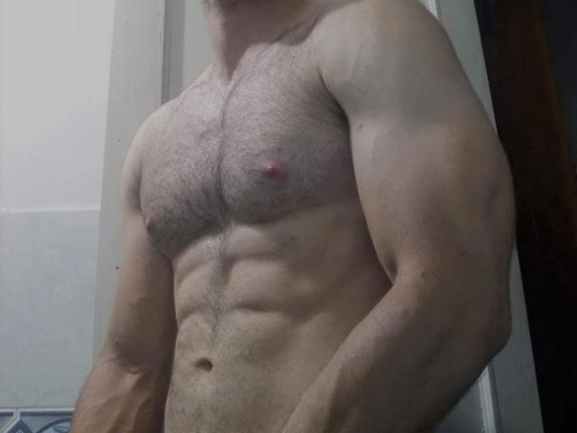 NachoMasterVidal's muscular, hairy chest with small, pink nipples and defined abs, showing a strong, tan-skinned upper body in a slightly angled, low-light photo.
