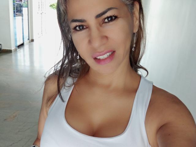 BrigiteWalsh has brown hair, tan skin, and is wearing a white tank top showing her tits. She's smiling with pink lipstick, long eyelashes, and dangling earrings.