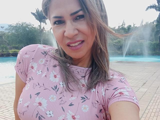 BrigiteWalsh with brown hair, pink floral shirt, smiling, blue pool, tan bricks, water spray, medium tits, light skin, pink lipstick, outdoors, sunny, mid-shot, selfie.