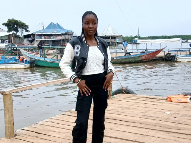 CutieLovelly, dark-skinned woman with small tits, wearing a black jacket, white lace top, black pants, standing on a wooden dock, hands on hips, looking at camera.