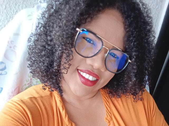 FionnaBigBan with curly black hair, wearing blue-tinted glasses, orange shirt, and red lipstick, smiling at the camera. Boob-free, no visible ass or pussy.