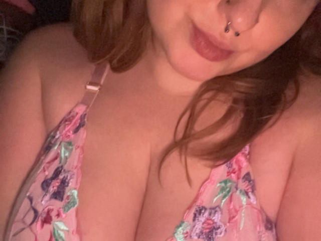 YourThickGingerGoddess with her nose ring, pouty lips, and pink floral lingerie, showing her big, fluffy tits and a peek of her cleavage in a slightly dim room.