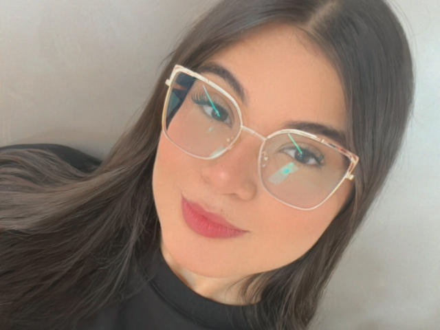 CamilaSofiia has long black hair, red lipstick, and is wearing clear-framed glasses with reflective blue eyes. She's smiling subtly, wearing a black turtleneck.