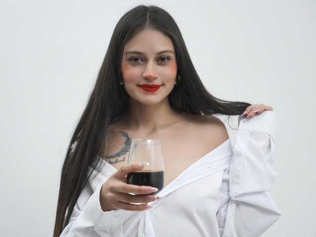 LizzaRose with long black hair, red lipstick, white off-shoulder shirt, holding a glass of red wine, visible tattoo on right shoulder, slight smile, white background.