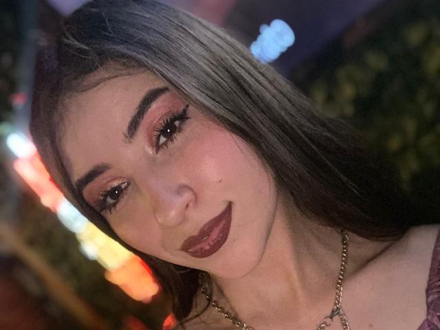 BrendaZambrano with long brown hair, full eyebrows, pink eyeshadow, thick eyelashes, dark lipstick, and a gold necklace, smiling at the camera in a dimly lit setting.