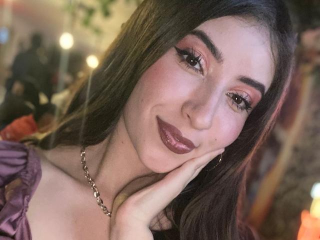 BrendaZambrano with long brown hair, pink eyeshadow, and dark lipstick, smiling with hand on cheek, wearing a purple off-shoulder top and silver chain necklace.