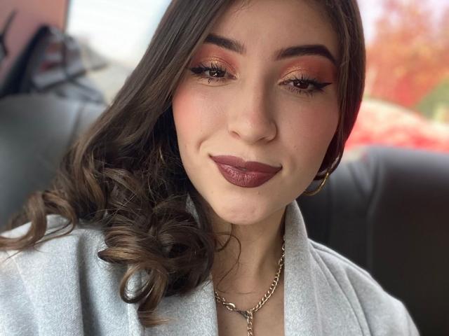 BrendaZambrano with curly brown hair, dark red lipstick, orange eyeshadow, and gold chain necklace, smiling in a gray top, seated on a black leather seat.