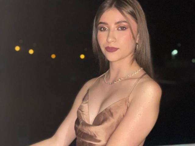 BrendaZambrano with long brown hair, wearing a brown satin dress, necklace, and dark red lipstick, has her tits slightly visible, standing in a dark, lit-up background.