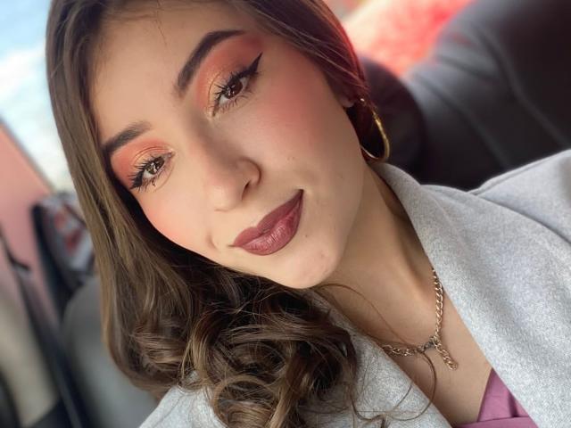 BrendaZambrano's face with pink and orange eyeshadow, thick eyebrows, long brown hair, glossy pink lipstick, gold hoop earrings, and a grey top, wearing a gold necklace.
