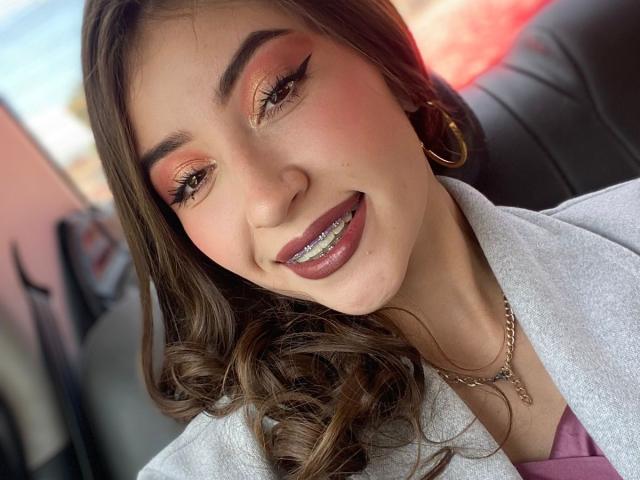 BrendaZambrano with brown wavy hair, pink eyeshadow, black eyeliner, smiling with her teeth showing, wearing a gray coat, gold hoop earrings, and a gold necklace.