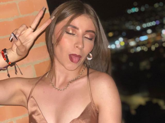 BrendaZambrano with brown hair, gold makeup, tongue out, pink lipstick, and a tan satin top, making a peace sign, wearing a chain necklace and red bracelet.