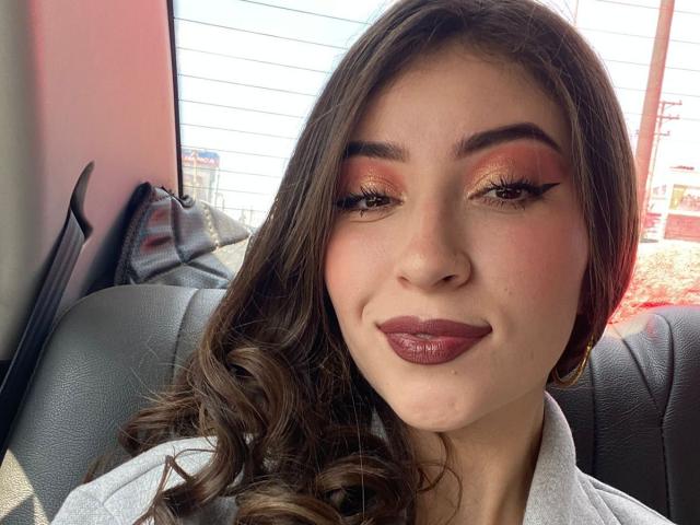 BrendaZambrano has shiny pink eyeshadow, thick black eyeliner, curled brown hair, and glossy dark red lipstick, smiling in a car with black leather seats.