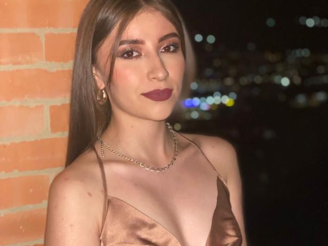 BrendaZambrano with long brown hair, wearing a shiny bronze dress, gold chain necklace, and dark red lipstick, standing against a brick wall at night. Boobs visible.