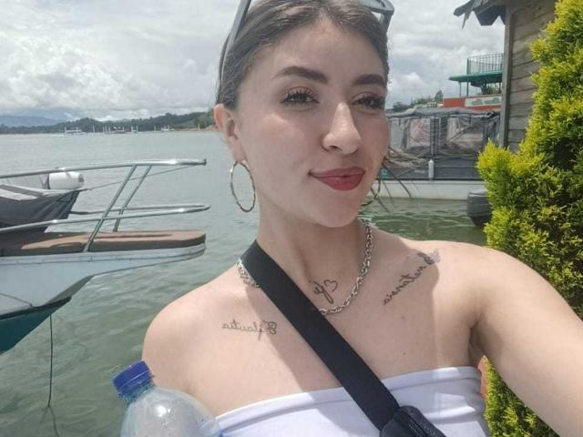 BrendaZambrano with small tits, tattoos on shoulders, white strapless top, red lipstick, hoop earrings, chain necklace, black strap, water bottle, boat dock, green bush, cloudy sky.