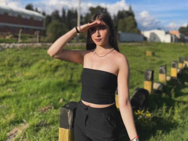 BrendaZambrano with black strapless top, black pants, fair skin, brown hair, hand on forehead, necklace, bracelet, standing in grassy field, wooden posts, sunny day, blue sky.