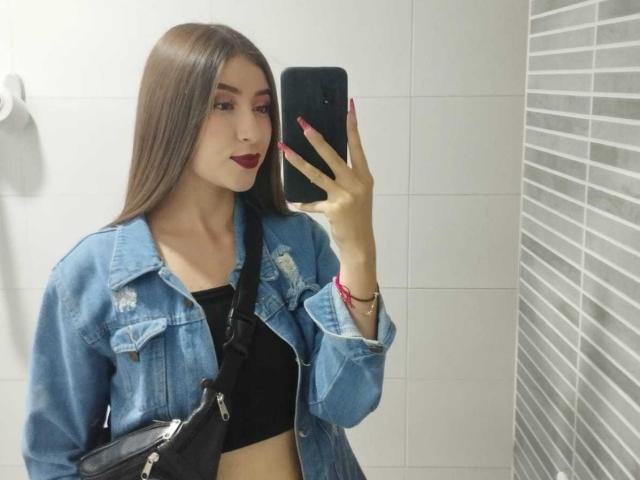 BrendaZambrano with long brown hair, red lipstick, wearing a denim jacket over a black crop top, holding a phone, mirror selfie, showing her tits, in a bathroom.