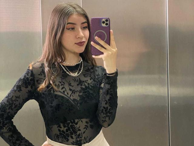 BrendaZambrano with brown hair, wearing a black lace top, white skirt, pearl necklace, and dark lipstick, taking a mirror selfie with a purple phone. Her tits are visible.