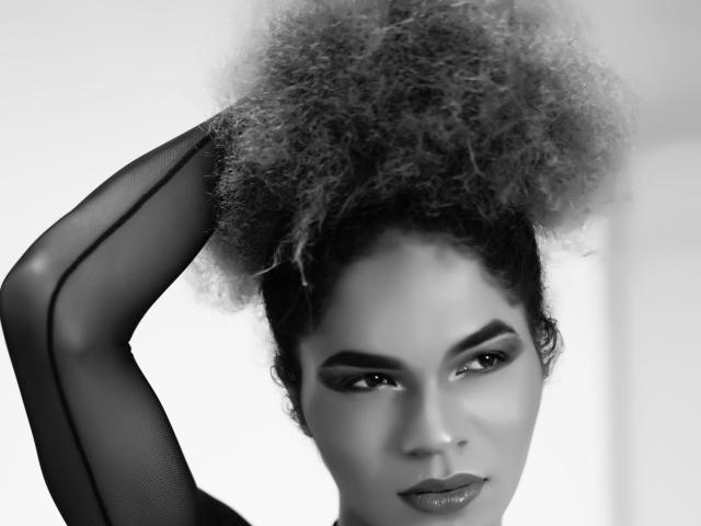 Black and white photo of Veennus with curly afro, wearing sheer black sleeves, raising arm, smoky eye makeup, and glossy lips, looking directly at camera with confident expression.