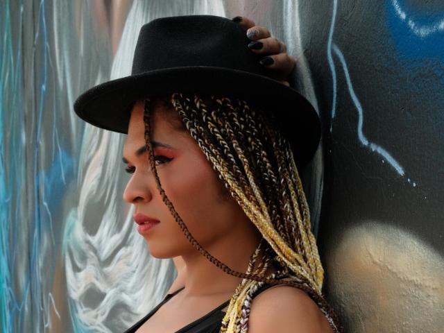Veennus with braided blonde and brown hair, wearing a black hat, black tank top, and red lipstick, leaning against a graffiti-covered wall, showing off her cute ass.
