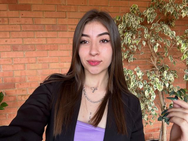 BrendaZambrano with long brown hair, purple top, black jacket, small tits, necklace, red lipstick, green nail polish, holding plant, brick wall background, smiling, medium skin, tattoo on chest.