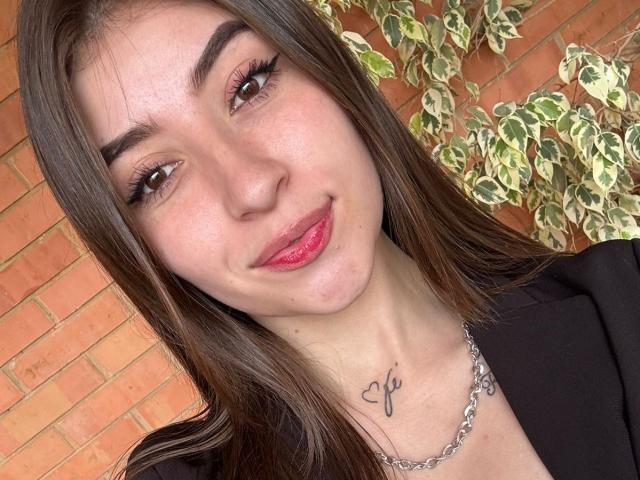 BrendaZambrano with long brown hair, brown eyes, and pink lipstick, smiling slightly, wearing a black top, necklace, and has a tattoo of a heart with an "F" below her chin.