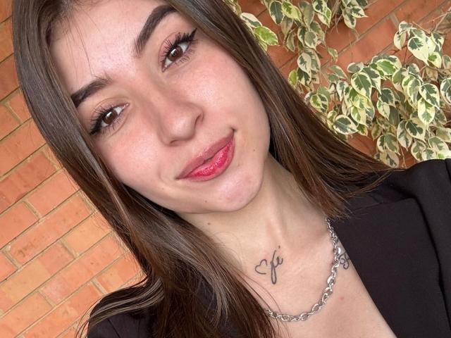 BrendaZambrano with brown hair, red lipstick, and a black top, showing a cross tattoo on her neck, wearing a chain necklace, smiling. Pupils are brown.