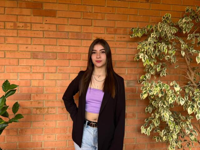 BrendaZambrano stands with hands on hips, wearing a black blazer over a purple tube top, blue jeans, and a necklace. Her brown hair is straight, lips are red.