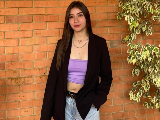 BrendaZambrano with long brown hair, wearing a black blazer, purple tube top, and blue jeans, standing against a brick wall, with a leafy plant on the right.