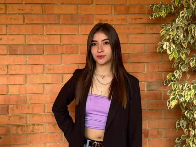 BrendaZambrano with long brown hair, purple tube top, black blazer, necklace, and hand on hip, showing her ass in tight jeans, standing in front of a brick wall.