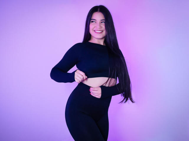 CamilaSofiia with long black hair, smiling, lifting her black crop top to reveal her bare, soft-ass, and wearing black high-waisted pants against a purple gradient background.