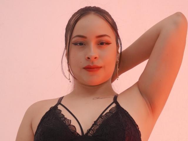 ValeriaJames with brown hair, black lace bra, arm raised, showing armpit, subtle tattoo on chest, eyeliner, and earrings, pinkish-orange light, confident expression.