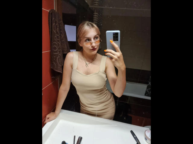 CatIlusionxx-hot with glasses, yellow nails, beige tank top, and cross necklace, taking a mirror selfie, showing her tits, standing in a bathroom with a white sink.