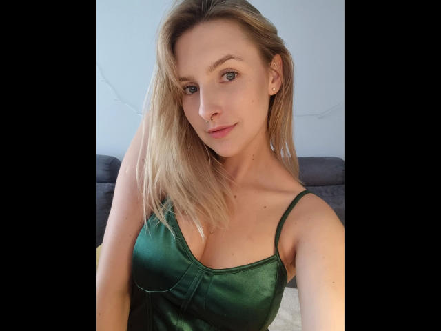 CatIlusionxx-hot with blonde hair, green eyes, and small tits in a green, shiny, spaghetti-strap top, smiling at the camera.
