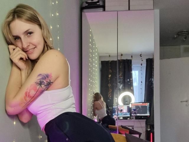 CatIlusionxx-hot with blonde hair, white tank top, black pants, and pink tattoo on arm, leaning against wall, smiling, reflecting in mirror, ring light on desk in background.