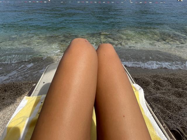 SandraSwweet's tan, smooth legs are on a yellow towel, with her knees touching, on a white boat, overlooking a blue-green ocean with gentle waves.