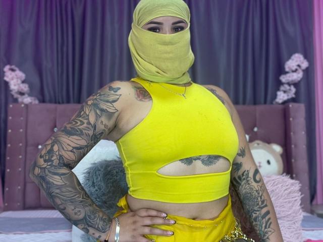 MalakCote mit gelber Maske, Ärmeltattoos, gelbem Crop-Top und gelber Hose, die Tattoos und ihre Brüste durch Ausschnitte zeigt, steht mit einer Hand auf der Hüfte und blickt in die Kamera..