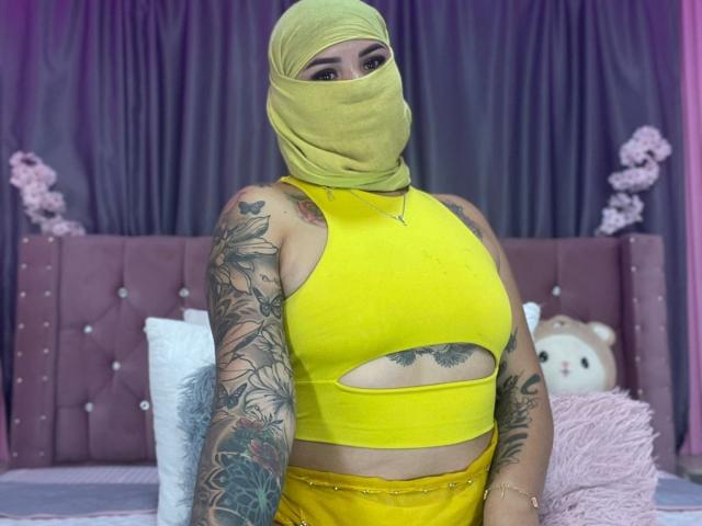 MalakCote with yellow mask, yellow cutout top, yellow pants, showing tits, tattoos on arms, sitting in pink room, plush toys in background, purple curtains, soft pink lighting.