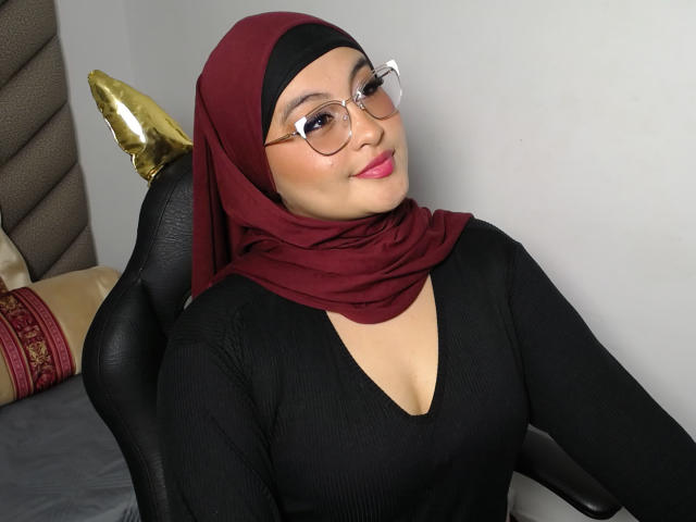 HelueJamil, con un hijab color borgogna, occhiali, una maglietta nera a coste e rossetto rosa, seduta su una sedia, sorride leggermente, con lo schienale dorato della sedia e un cuscino con motivi rossi sullo sfondo..