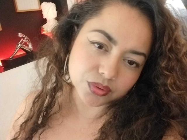 Lalitzfet with curly brown hair, medium skin, and pink lipstick, pouting slightly, wearing hoop earrings, looking at the camera. Boobs partially visible. Soft, natural light.