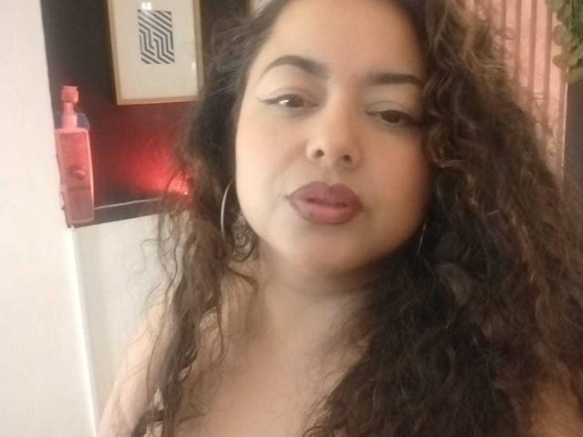 Lalitzfet ha i capelli castano ricci, la pelle abbronzata e ha le labbra arricciate con un rossetto rosa. Indossa degli orecchini a cerchio e guarda leggermente di lato con un'espressione rilassata..