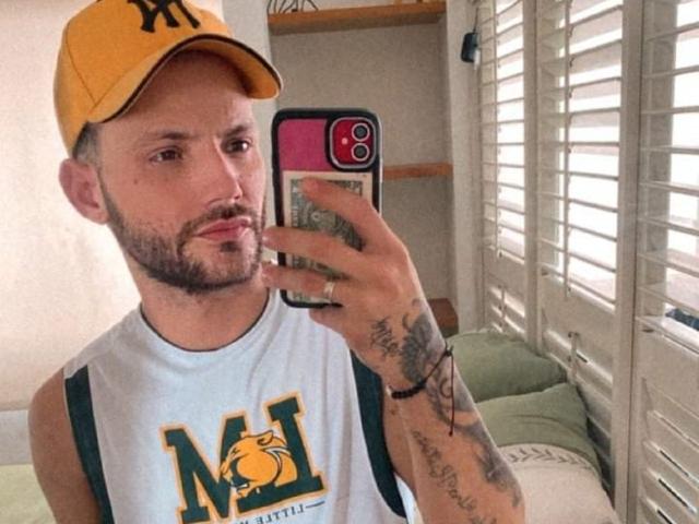 BladeBigCock69, bearded dude with tattoos, wearing yellow cap, white tank top, taking selfie in mirror, holding phone with pink and black case, green and white shirt.