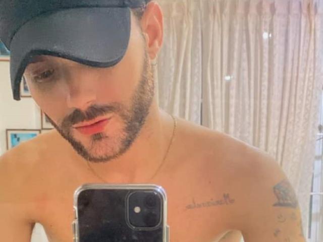 BladeBigCock69 with a black cap, beard, and tattoos on his shoulder and chest, taking a mirror selfie with a phone, shirtless, and looking down.