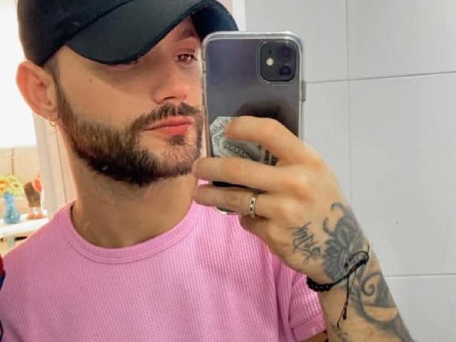 BladeBigCock69, a bearded dude with tattoos on his hand and forearm, wearing a pink ribbed shirt and black cap, takes a mirror selfie with a smartphone.