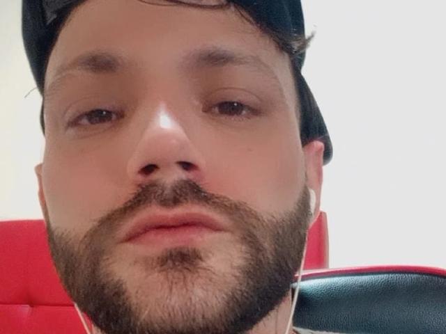 BladeBigCock69 has a medium beard, brown eyes, and is wearing a black cap with white earbuds in. Red chair is visible in the background.