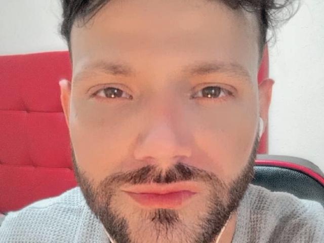BladeBigCock69 has a bearded face with short dark hair, brown eyes, and a slight mustache, wearing a gray sweater, sitting in a red chair.