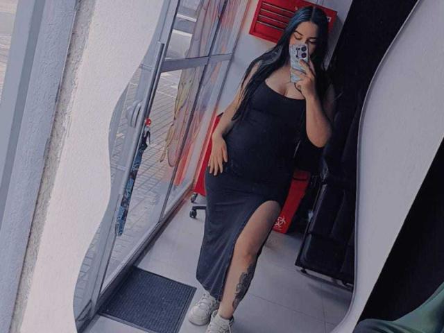 Aurora, com longos cabelos pretos, vestindo um vestido preto com um ombro descoberto, com uma fenda na coxa, tênis brancos, segurando um celular, com uma tatuagem visível na perna, em pé na porta, tirando uma selfie no espelho..