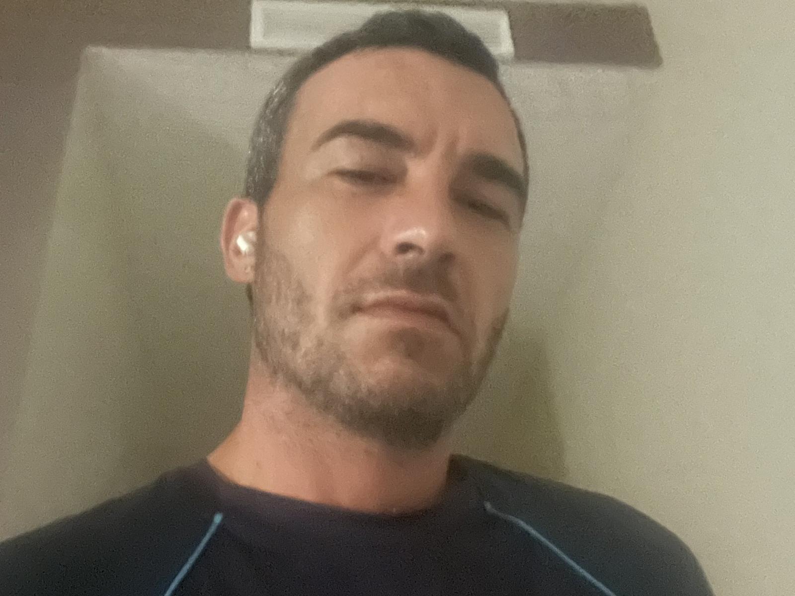 ThiboChaud heeft kort donker haar, een baard, en draagt witte oordopjes. Hij heeft een licht geïrriteerde uitdrukking. Hij draagt een donker shirt met blauwe naden..