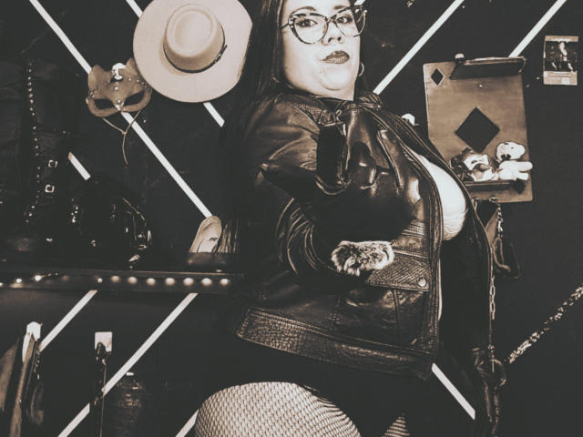 LexyDom with glasses, leather jacket, and fishnet pants, holding a whip, hat in hand, exposing her big tits, black lipstick, and a confident, sexy look. Sepia-toned.