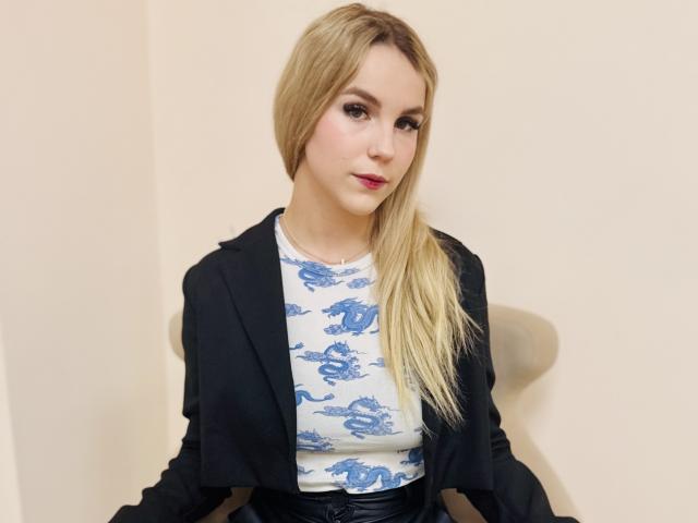 PaulaSpency with blonde hair, blue dragon shirt, black blazer, and red lipstick, sitting, looking at camera, showing her tits slightly, in a plain room.